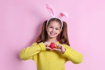 Studio portrait of smiling young girl wearing traditional bunny ears headband for easter. Brunette female with pigtails holding traditional colored eggs over pink background. Close up, copy space.
