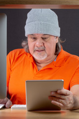 Man working on computer at his desk. He is in his 50s. He is wearing a t-shirt and a hood. He has long hair. This is a studio image. He is looking at the tablet screen.