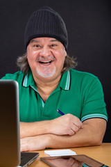 Man poses working on laptop at his desk. He is in his 50s. He is wearing a t-shirt and a hood. He...