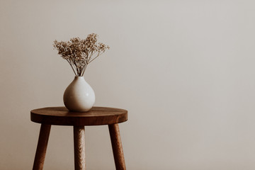 White ceramic vase on a natural brown wooden stool with white dried flowers in an empty room