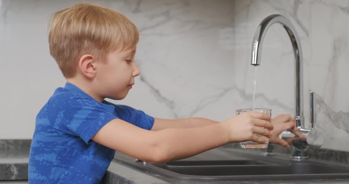Child takes a clear glass, fills it with fresh water and drinks it. Blond boy goes to the kitchen tap opens it pours water and drinks.