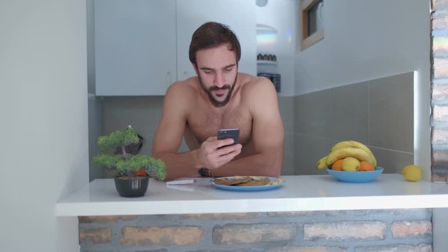 Shirtless, smiling, fit man scrolling on his mobile phone while leaning on the kitchen counter with protein pancakes on. Pan shoot from left to right