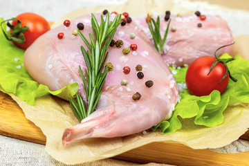 Raw uncooked pink rabbit leg meat with green rosemary and fresh vegetables on light wooden cutting board background.