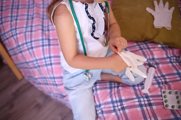 Fototapeta premium Little attractive Caucasian girl puts on medical gloves plays a doctor with a stethoscope and smiles. Health Concept. Pandemic. Quarantine. Insulation. Stor coronavirus 2019-ncov