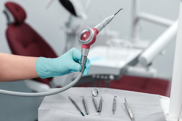 Female hand of dentist in blue gloves holding sandblasting tool, blurred background of clinic with...