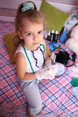 Caucasian little serious girl in a medicalg gloves with stethoscope at home playing doctor with a syringe for injections prepare to treat a bear. Little therapist. Coronavirus 2019-ncov. Home insulat