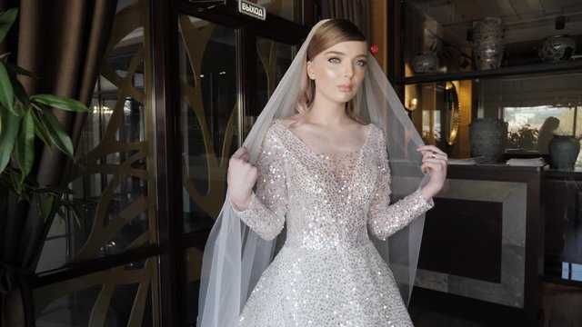 attractive blonde bride poses for photo shoot holding long veil in hands against restaurant decorations slow motion