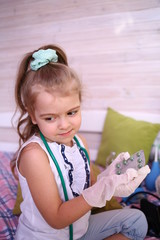 Quarantined fun. Caucasian cute little girl in a medical gloves at home playing doctor with a bear, give pills, treat. Quarantine protection against coronavirus 2019-ncov. Home insulation