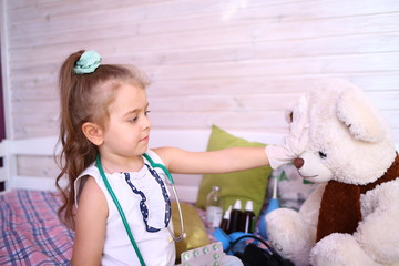 Quarantined fun. Caucasian cute little girl in a medical gloves at home playing doctor with a bear, 
checks forehead and temperature. Quarantine, coronavirus 2019-ncov. Home insulation