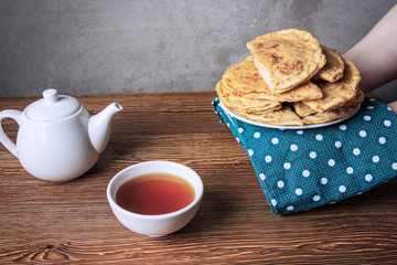 Hands holding a bowl of traditional chebureks. On the table is a white teapot and a bowl filled with hot tea