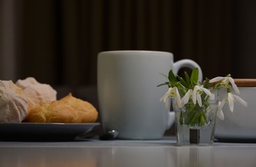 Tea and plate with eclairs and marshmallows on a transparent table. Vase with snowdrops.