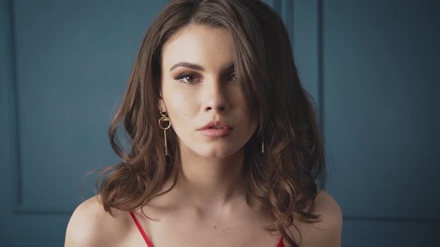 Beautiful brunette woman lady in a silk red dress evening make-up leans against a blue wall to bask in a luxurious life glamorous model style fashion jewelry tender skin youths attractive sexy female.
