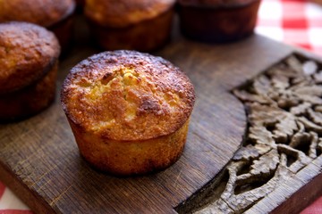 Single muffin on a wooden carved board with other muffins in the background.  A checkered red and white table clothes peaks through