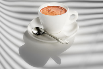White cup of coffee on white background with shadows from the window blinds. Best start to the day. Copy space.