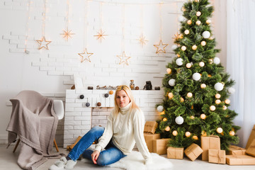 Christmas fashion photo of beautiful girl with blond hair in cozy clothes posing near New Year tree with presents