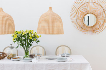 Grey plates, vine glasses and yellow flowers in vase in trendy dining room interior with wicker chandeliers