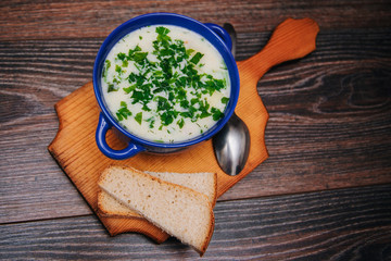 A blue cup with cheese, vegetable soup stands on a cutting, wooden board. Next to the soup with herbs, two slices of bread