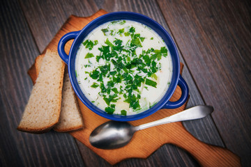 A blue cup with cheese, vegetable soup stands on a cutting, wooden board. Next to the soup with herbs, two slices of bread