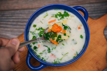 A blue cup with cheese, vegetable soup stands on a cutting, wooden board. Hand with a spoon scoops up soup with herbs
