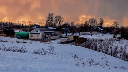 Beautiful winter landscape with a small village on a hill against the backdrop of a sunny sunset in the cloudy sky. Russia, Arkhangelsk region