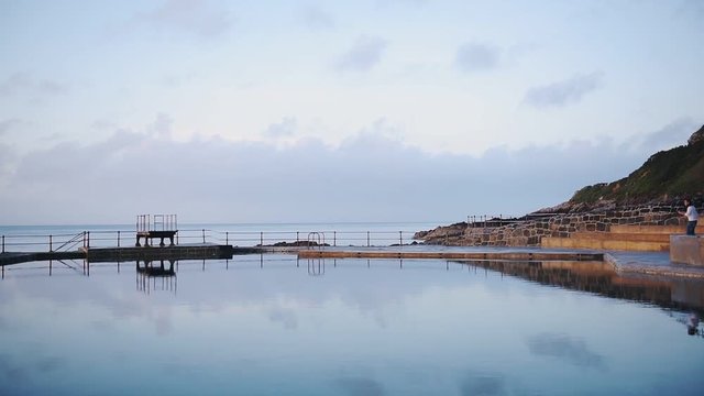 A Man Walking To The Deck Of The Swimming Pool To Capture The Beauty Of Guernsey Waters. -wide shot