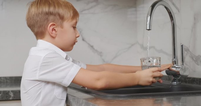Child takes a clear glass, fills it with fresh water and drinks it. Blond boy goes to the kitchen tap opens it pours water and drinks.