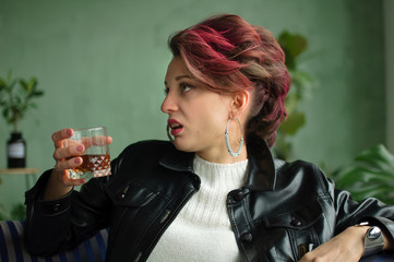 Young sad woman with dark pink hair with glam rock style look is drinking whisky from a glass with abomination grimace on the face, female alcoholism, alcohol addiction