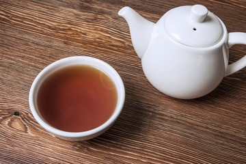 A bowl filled with hot tea next to a white teapot on a wooden table, top view