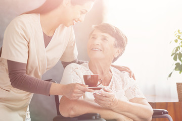 Smiling nurse giving hot tea to happy elderly lady in a wheelchair at nursing home
