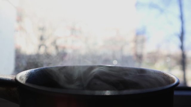 Steam rises while cooking in a pan. Open a small pan with steam. The stewpan stands on the windowsill near the window and cools down. Cook at home