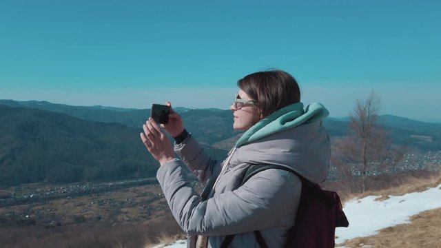Female hiker taking photos of beautiful mountains on smartphone on a bright sunny day on top of a mountain. 