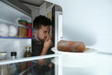 Young man feeling bad smell of spoiled sausage in refrigerator, view from inside