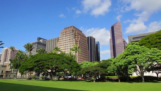 A town of modern high-rise buildings rise up to the blue sky and white clouds of Honolulu on the island of Oahu as lush, full trees rest at their base.