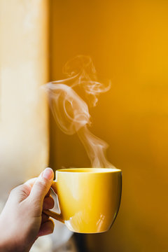 Yellow cup with hot steaming tea drink in hand near window with bright yellow wall background