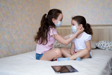 little girls, sisters in protective masks adjust each other's masks while at home in self-isolation on the bed