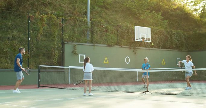 Happy Caucasian family playing tennis match on sport court on sunny summer day. Parents with kids play sport game outdoor. Children with mother and father having fun on weekend.