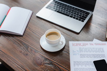 High angle view of cup of coffee near copybook, laptop and smartphone on papers on table in office