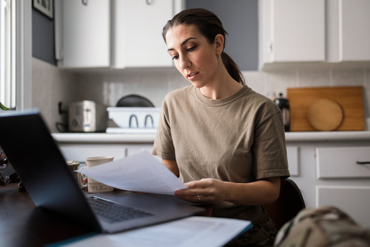 Female soldier reading paperwork at laptop in kitchen