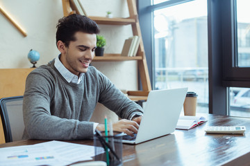 Selective focus of businessman smiling and working with laptop at table in office