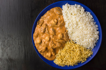 Brazilian tenderloin stroganoff with rice and potato sticks in a blue plate isolated in dark wood background  seen from above