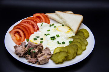 Delicious fresh tuna with eggs, bread, tomatoes, cucmbers with green onion at the morning. Healthy and diet breakfast with tuna, eggs and vegetables on the white plate against dark background