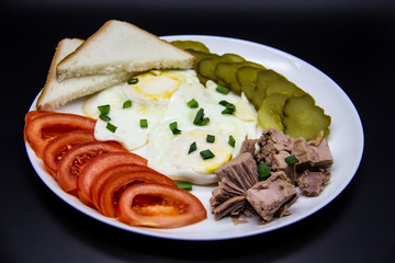 Delicious fresh tuna with eggs, bread, tomatoes, cucmbers with green onion at the morning. Healthy and diet breakfast with tuna, eggs and vegetables on the white plate against dark background