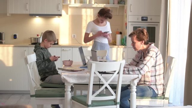 Mother, son and daughter looking, scrolling at their digital devices, smartphone and laptop in the kitchen separately, no communication