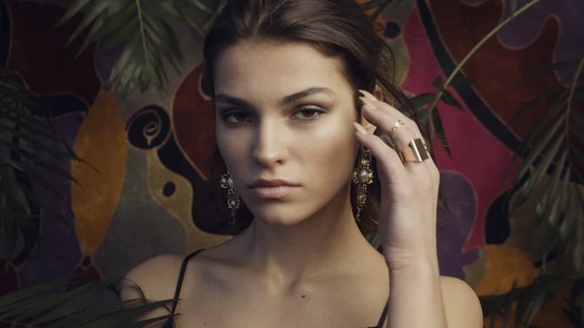 Tanned beautiful girl model with large gold earrings on a motley background among tropical leaves.