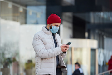 A caucasian man wearing a medical face mask to avoid the spread coronavirus (COVID-19). A guy with a surgical mask on the face using a smartphone in the center of the city.