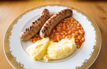Two large fried sausages with baked beans and mashed potato on a white plate with a gold colour trim on a wooden table lit by window natural light.
