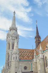 Fototapeta na wymiar The spire of the famous Matthias Church in Budapest, Hungary. Roman Catholic church built in the Gothic style. Orange colored tile roof. Blue sky and white clouds above. Vertical photo with filter