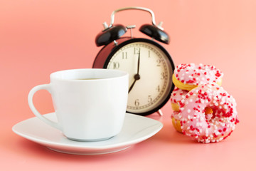 White mug on a saucer with black coffee, near a retro black alarm clock and donuts on a pink background. Breakfast time. Place for text