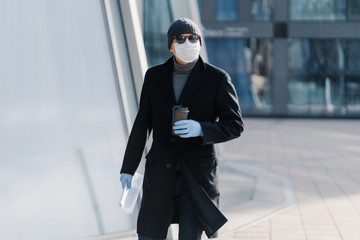 Outdoor shot of young man in black coat, sterile medical mask and protective rubber gloves, drinks takeaway coffee, holds papers, has to go working poses at empty street during quarantine, coronavirus