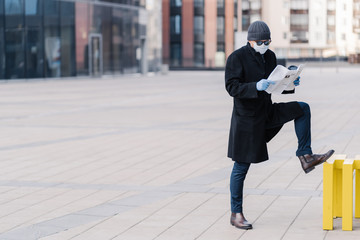 Sick man with flu wears medical mask to protect spreading infectious disease, dressed in coat, stands at street near office building, reads newspaper, finds out news about coronavirus pandemic